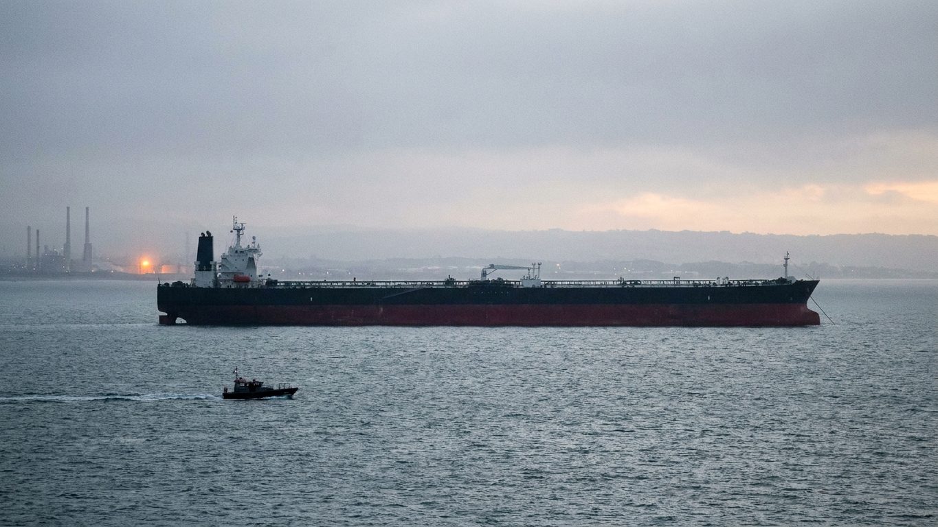 A crude carrier lying at anchor in ballast off a refinery terminal at dawn, a pilot launch crossing the foreground